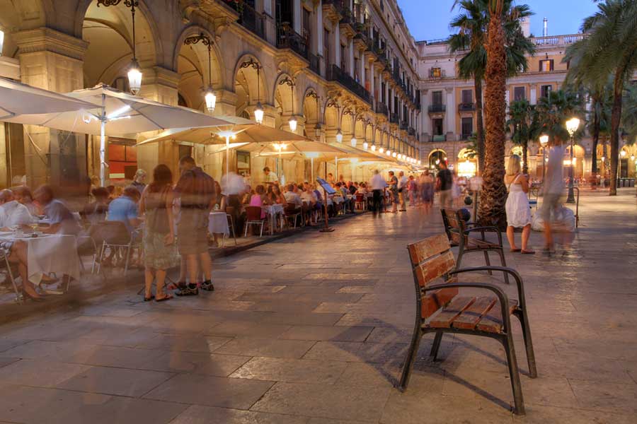 People sitting at outdoor tables on an old square at night, lit by streetlamps