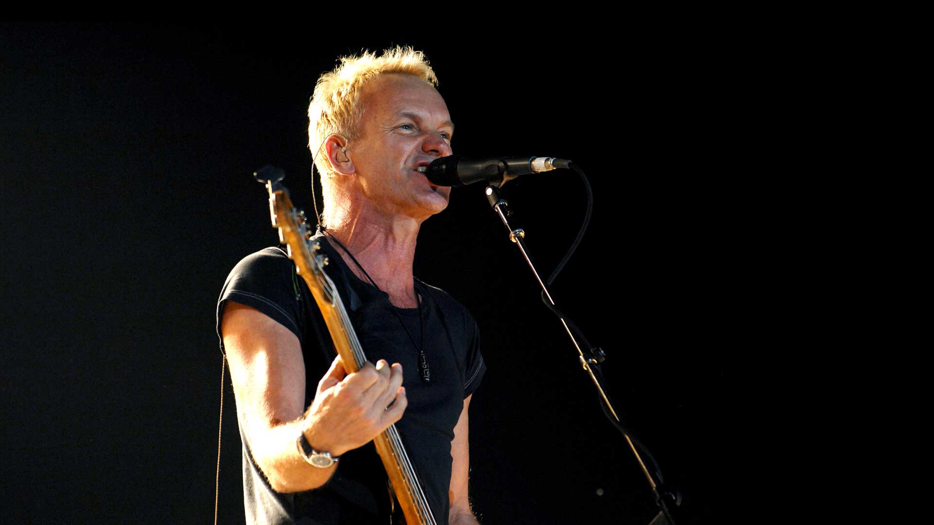 Musician Sting in a black t-shirt playing guitar and singing with a microphone in front of a black background