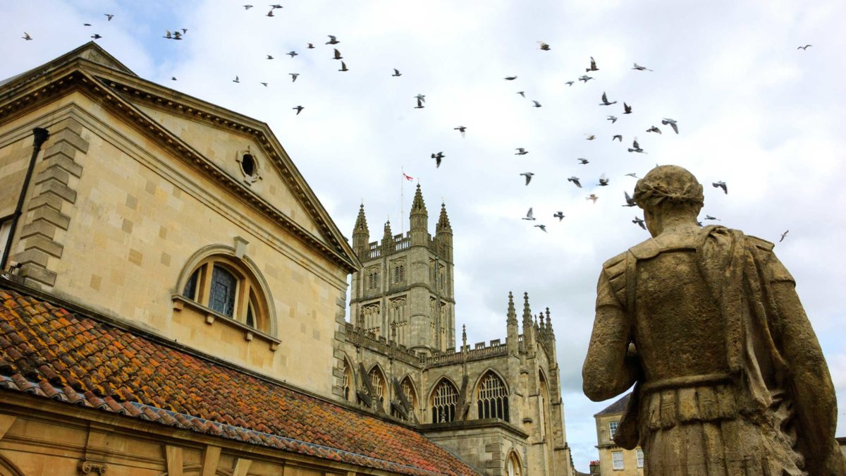 Stone statue of the roman in Antique Roman Baths complex, flying birds in sky and Abbey Cathedral at background.