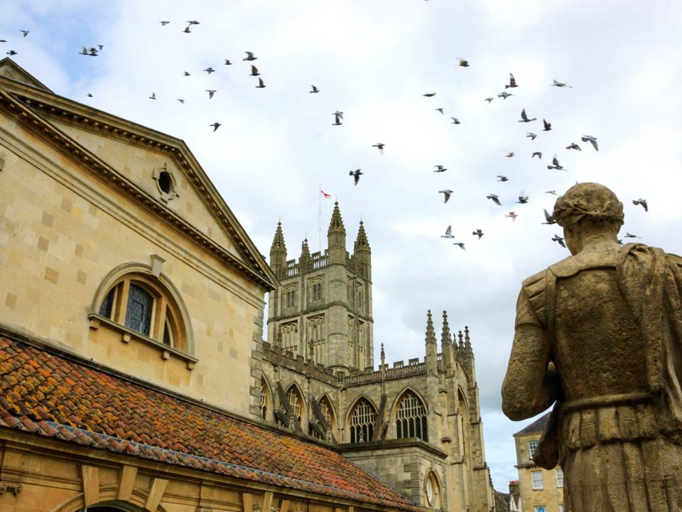 Stone statue of the roman in Antique Roman Baths complex, flying birds in sky and Abbey Cathedral at background.