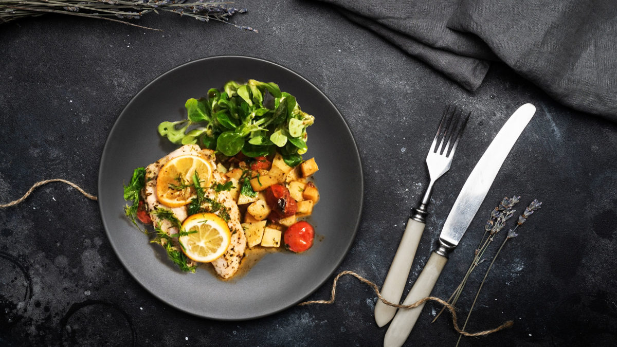 Baked Lemon & Herb Fish With Cherry Tomatoes on black plate with black dinner table and cutlery set